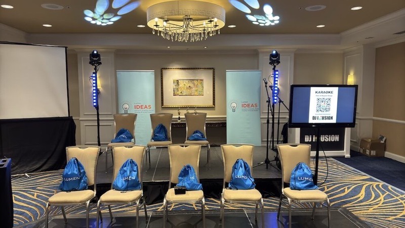 Stage with a row of chairs under a chandelier, two blue lighting totems, and a TV showing a karaoke song-request QR code by the DJ Illusion booth