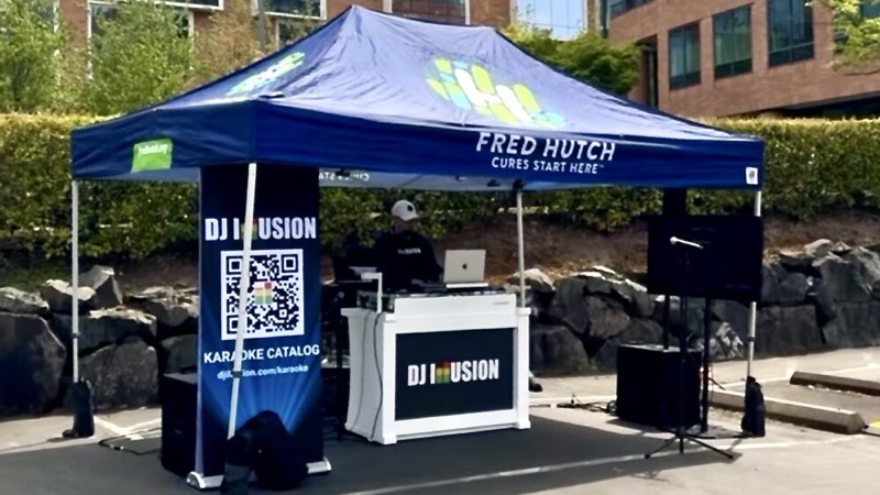 DJ in a cap at a white DJ Illusion booth under a pop-up canopy outdoors, with a karaoke catalog QR banner beside the booth
