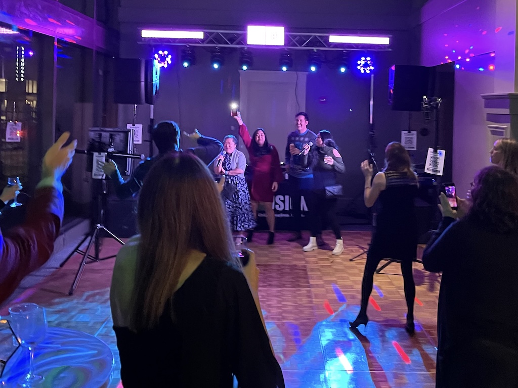 Guests singing karaoke into wireless microphones with hands raised as the crowd cheers