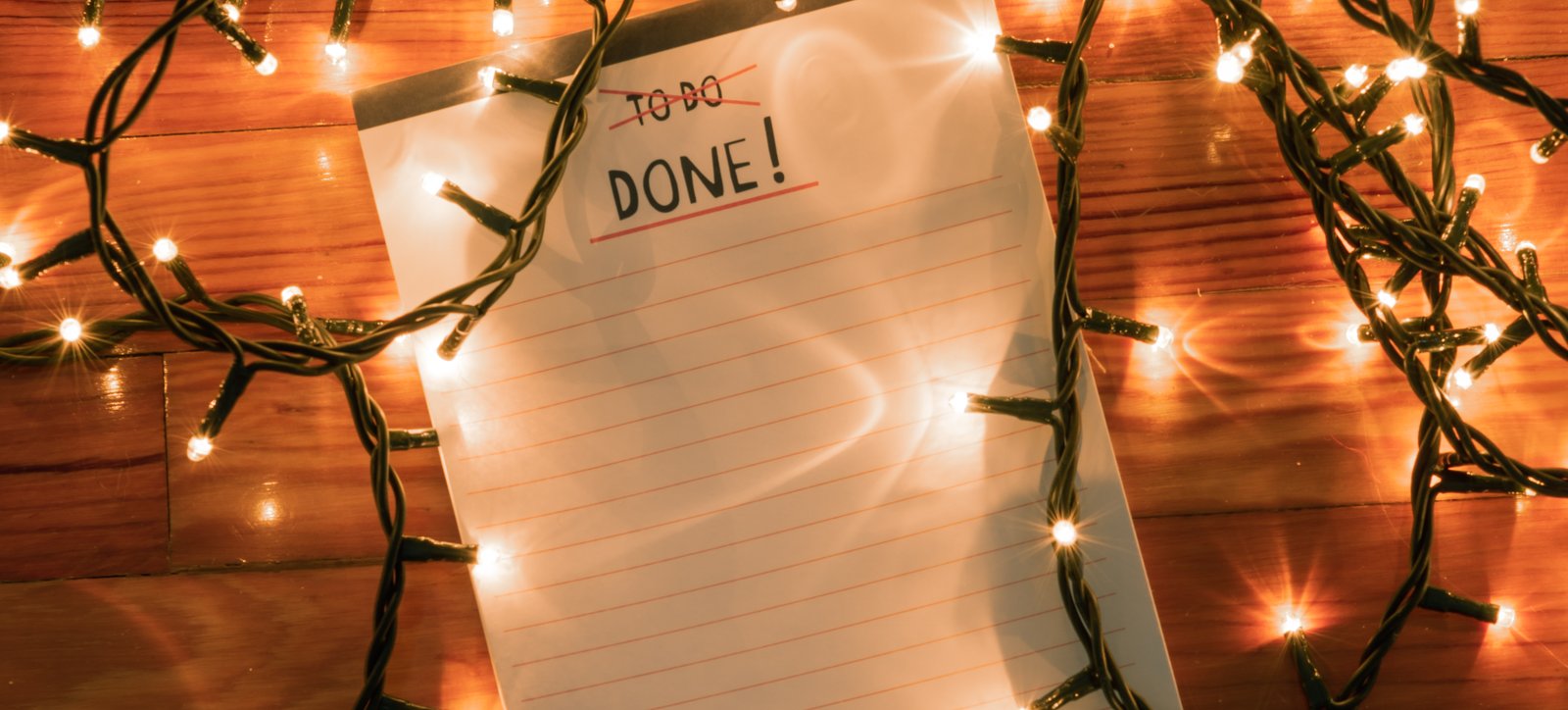 A notepad with "To do" crossed out and "Done!" written above blank lines, surrounded by warm string lights on a wooden floor