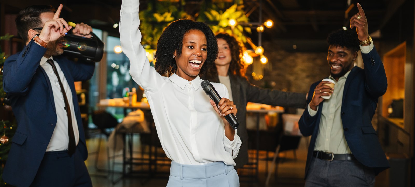 A woman sings into a handheld microphone, arm raised, as coworkers in business attire dance behind her at an office holiday party