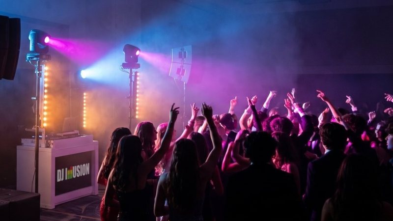Students with hands raised on a packed school dance floor beside the DJ Illusion booth, lit by blue and magenta moving lights through haze