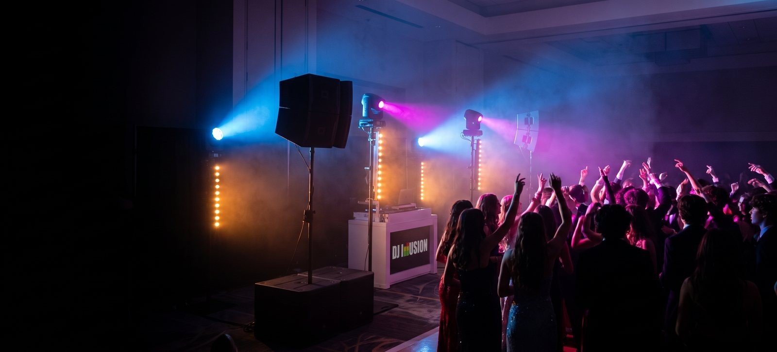 Students with hands raised on a packed school dance floor in front of the DJ Illusion booth, lit by blue and magenta moving lights through haze