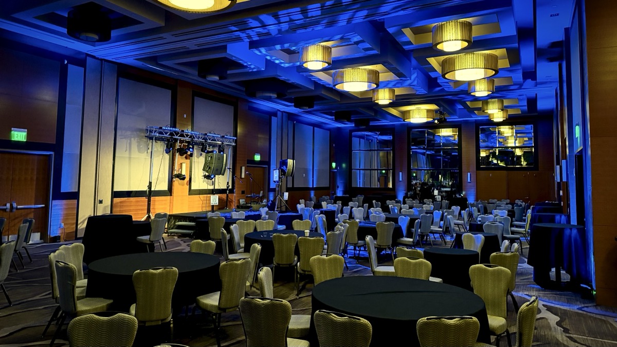 A hotel ballroom of banquet tables washed in blue uplighting with a lighting truss beside the dance floor